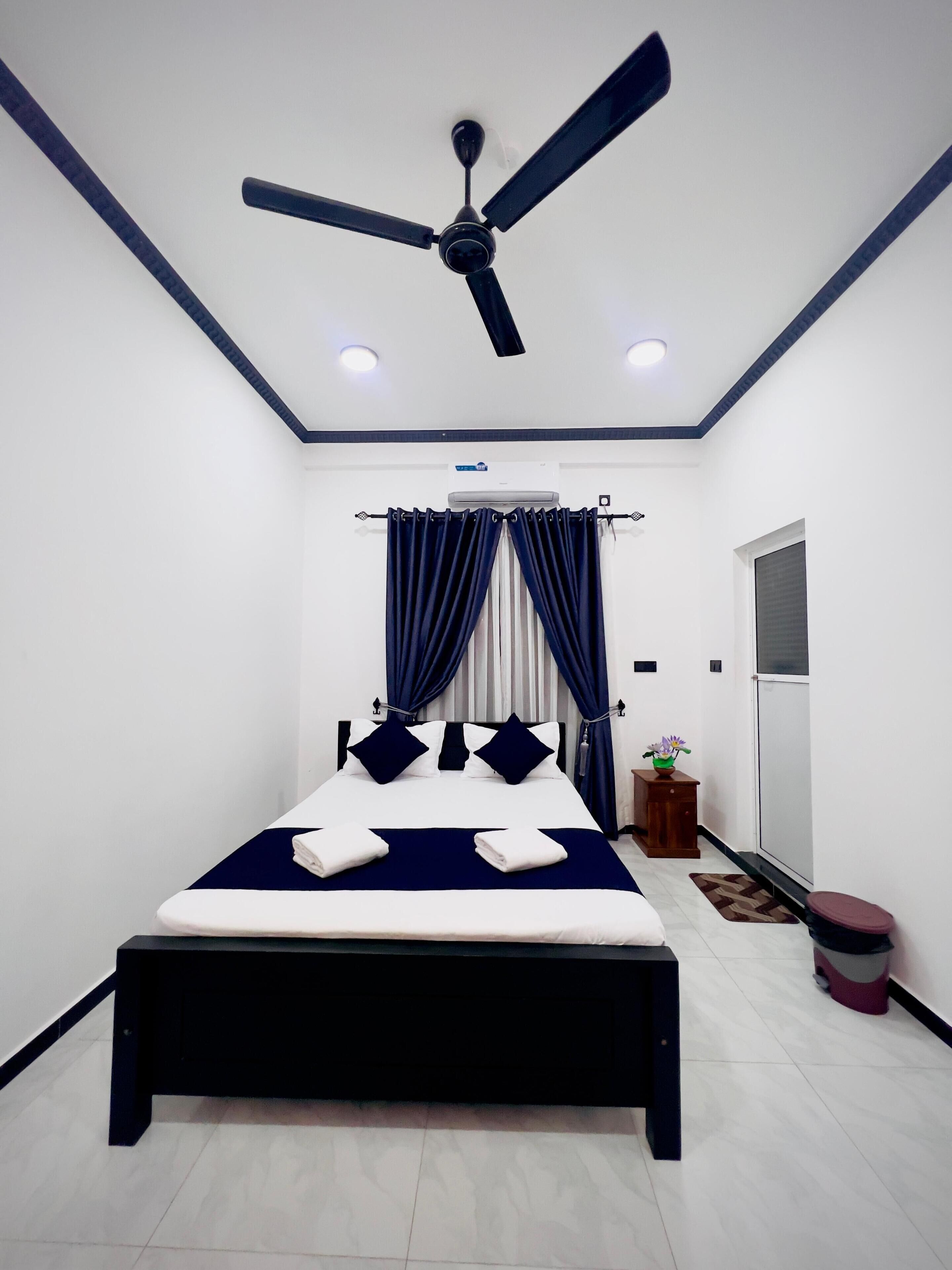 A bedroom with a bed, ceiling fan, air conditioning unit, and a small table with a plant.