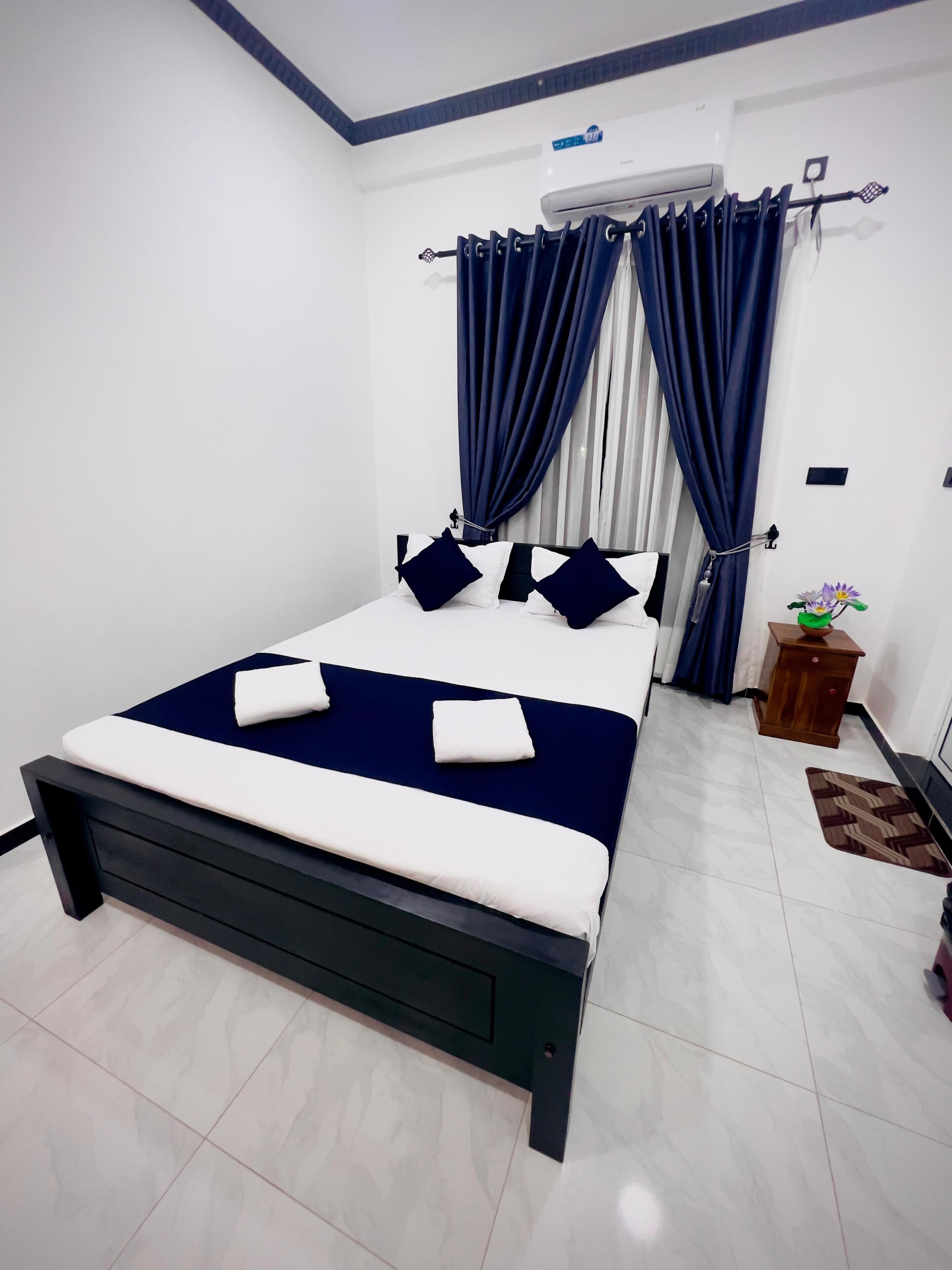 A bedroom with a bed, curtains, an air conditioning unit, a small wooden nightstand, and a floor mat.