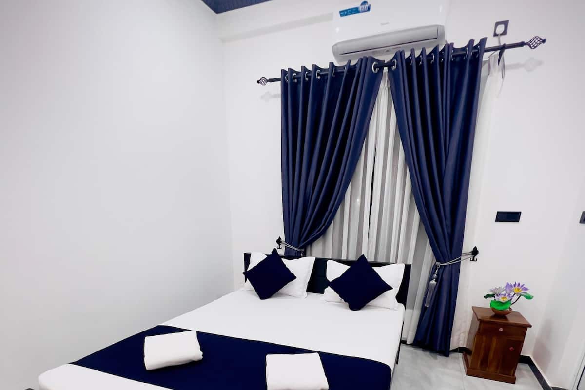 Un dormitorio con cama, cortinas, unidad de aire acondicionado, una mesita de noche de madera y un felpudo.
