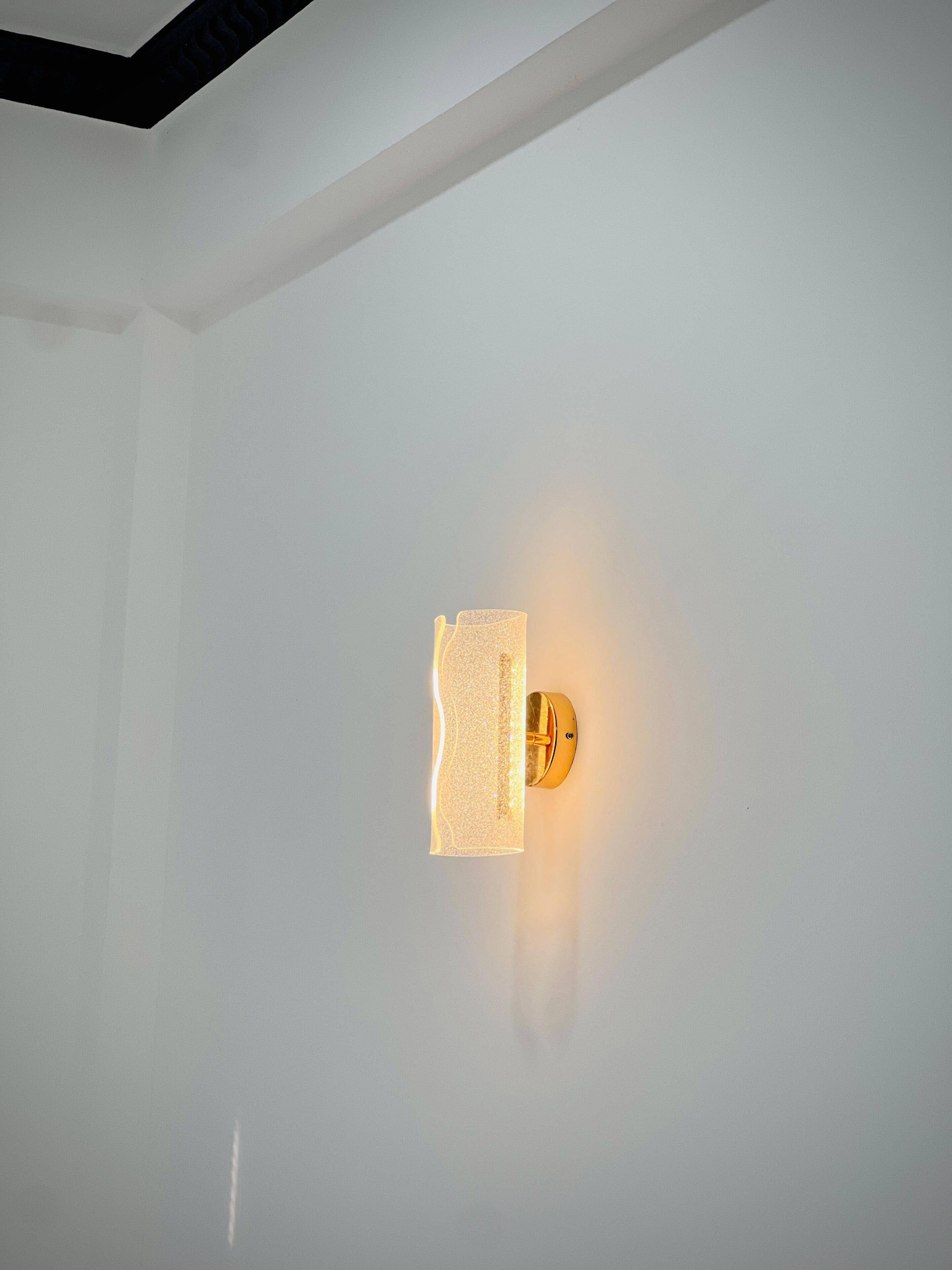 A wall-mounted light fixture with a textured glass panel and a gold-colored base.