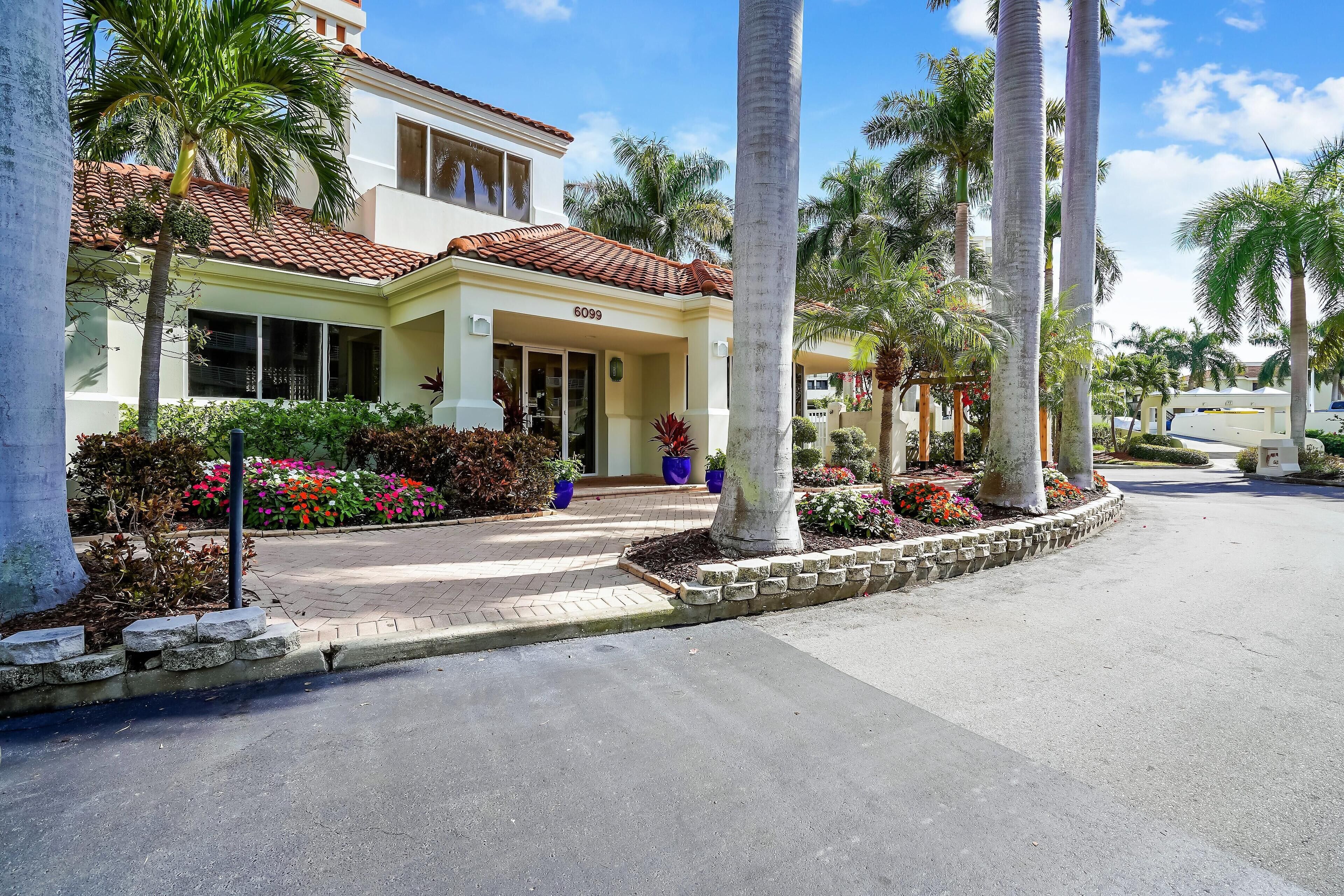 A two-story house with a number 6099 on the front door, surrounded by palm trees and colorful flower beds.