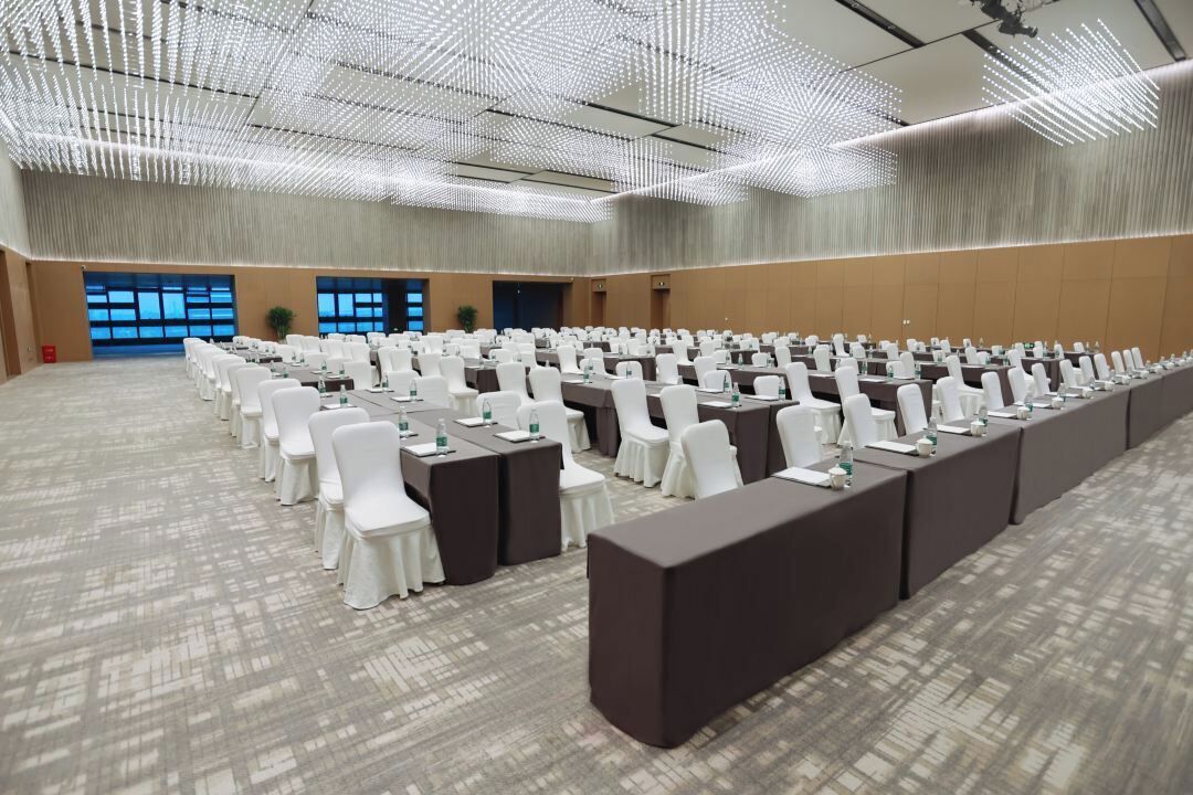 A large conference room with rows of white chairs and tables, a patterned carpet, and a ceiling with a unique light fixture.