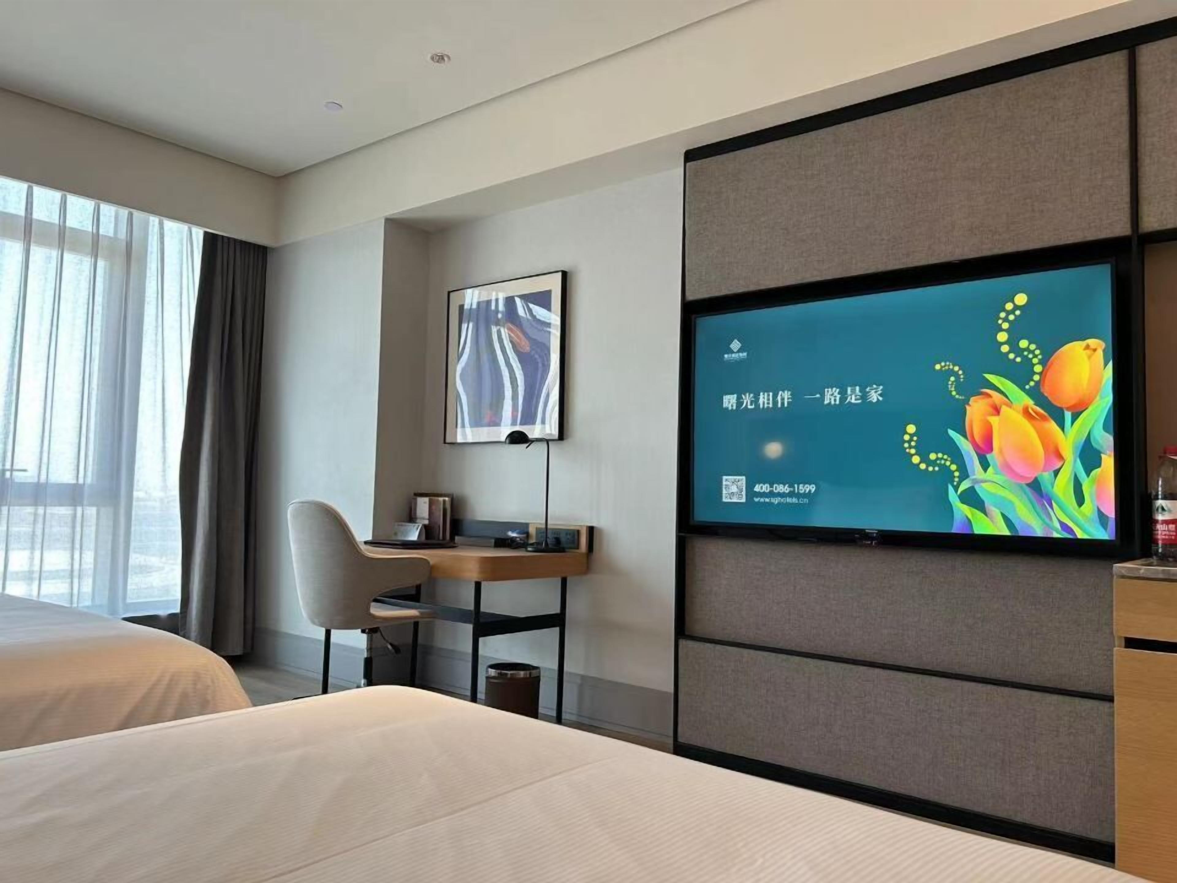 Quarto de hotel com cama, escrivaninha com cadeira, televisão na parede e janela com cortinas.