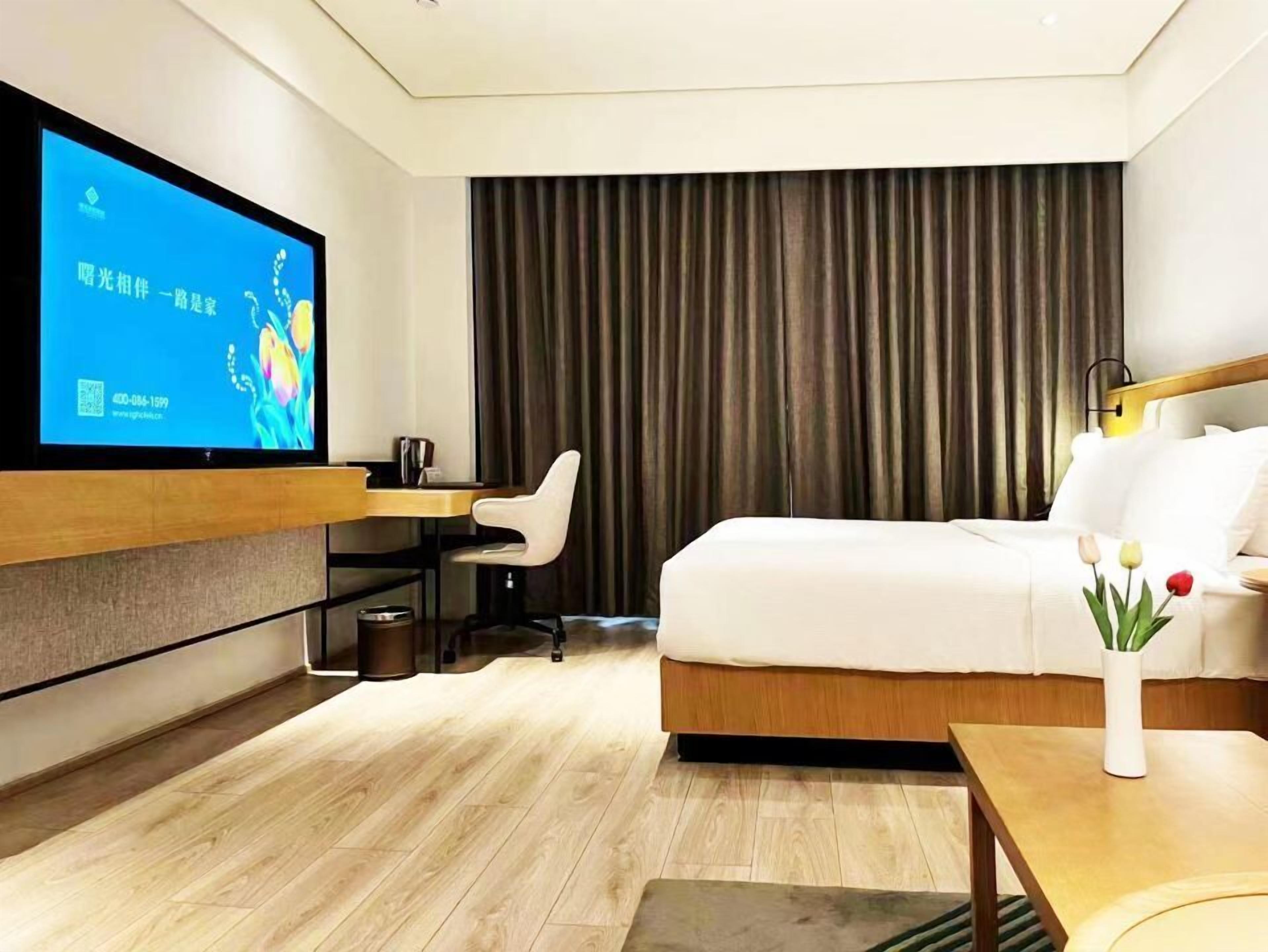 A modern hotel room with a flat-screen TV, a wooden desk, a comfortable chair, a bed, and a small table with flowers.