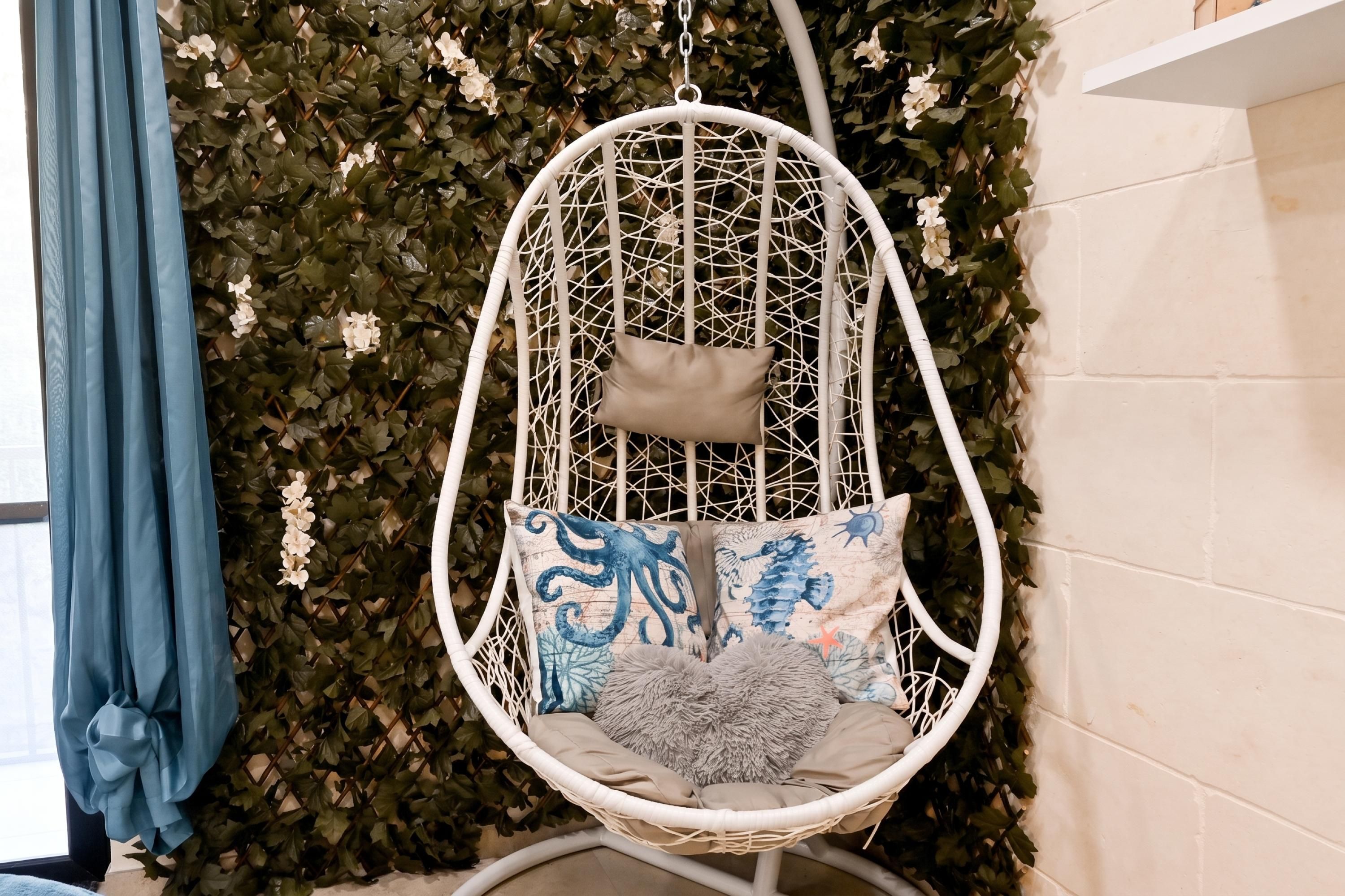 Une chaise suspendue blanche, dotée d’un coussin et d’un oreiller, est placée contre un mur recouvert de feuillage verdoyant.