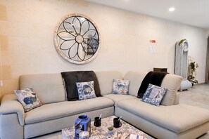 A living room with a beige sectional sofa, decorative pillows, a wall-mounted circular mirror, and a small table with a coffee mug and a candle. - Xlendi bay Wonderfull 3 bdr Apart Gozo (Munxar)