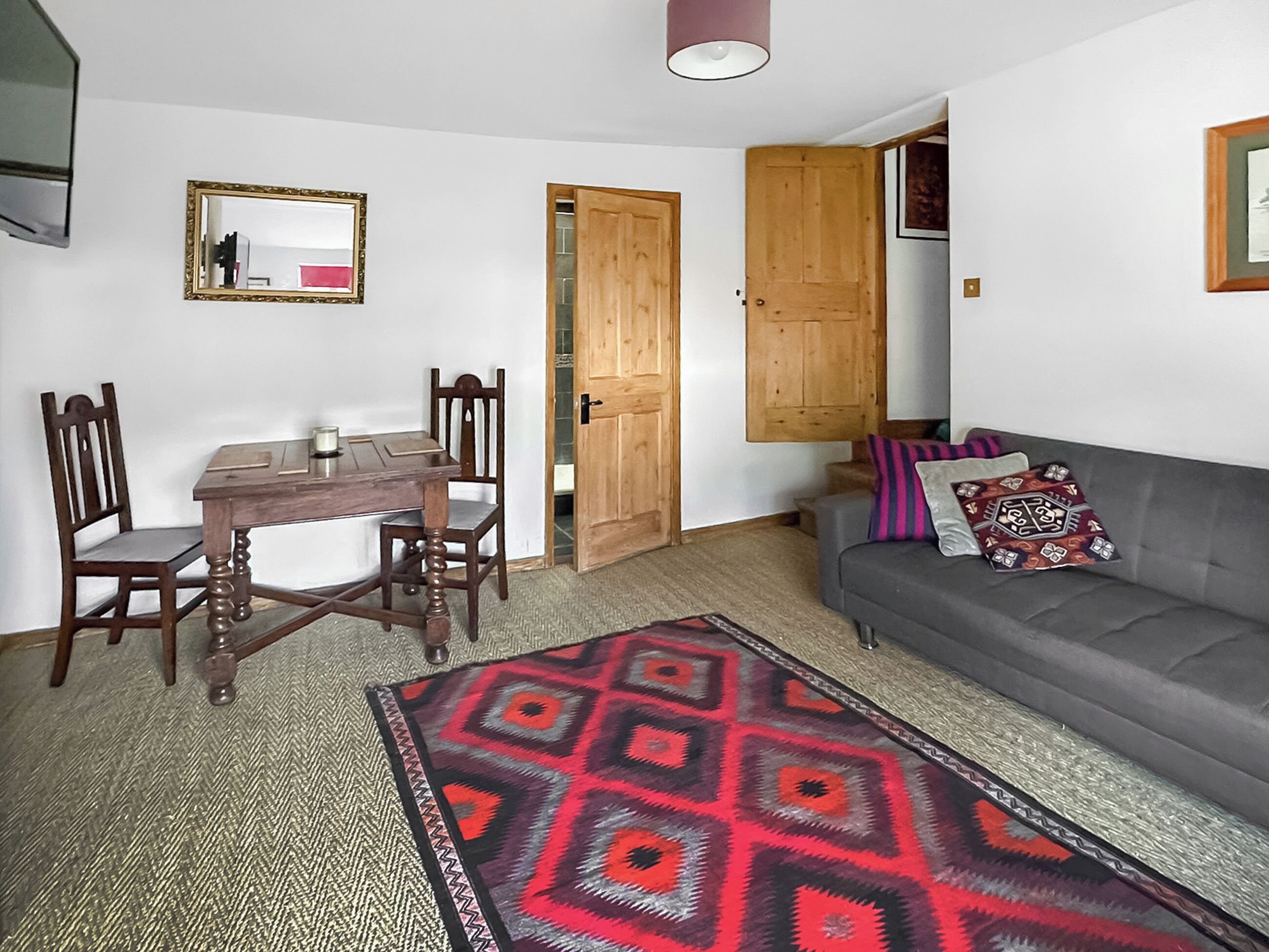 A room with a dining table, chairs, a sofa, a patterned rug, a mirror, and wooden doors.