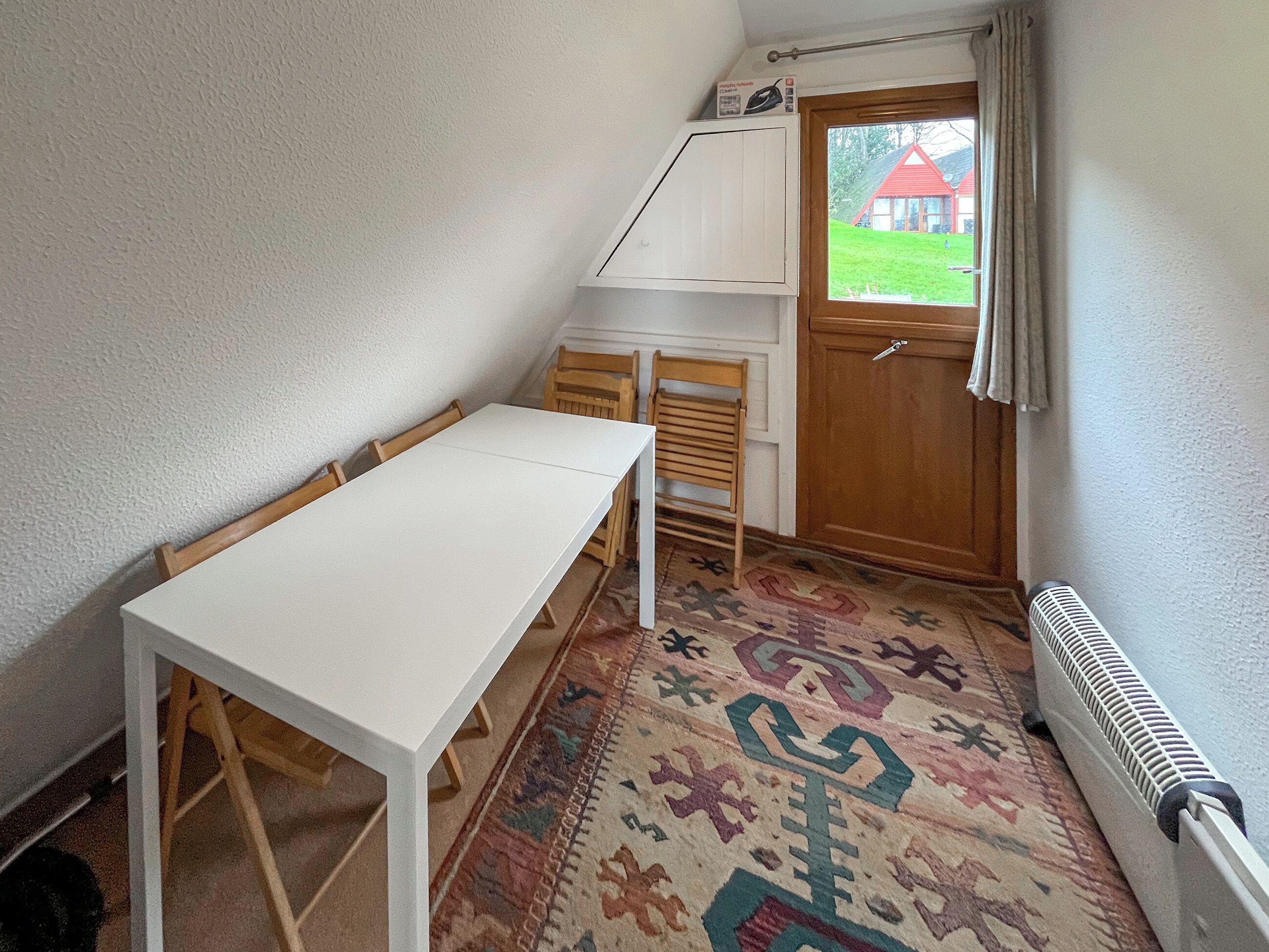 A small, cozy room with a white table, two wooden chairs, a patterned rug, a radiator, and a door leading to a garden with a red building.