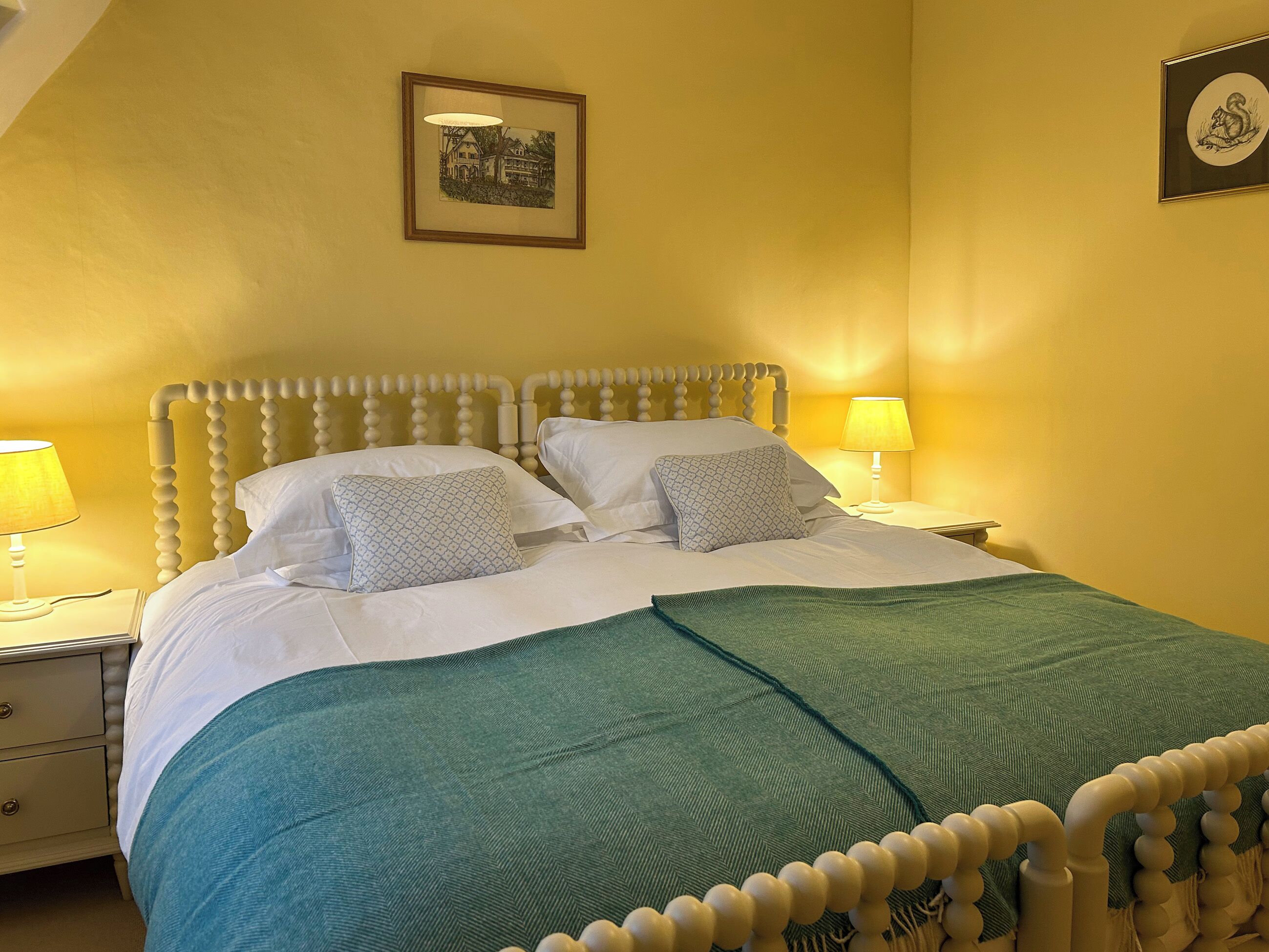 Een bed met een houten hoofdbord, twee bedlampen, een groene plaid en een lijst met een foto aan de muur.
