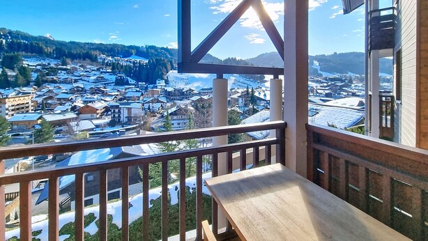 A balcony with a wooden bench, offering a view of a snow-covered town and mountains. - Okoume-Apartment-4pers-Proche center (Les Gets)