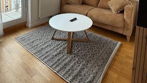 Una sala de estar con sofá, una mesa de centro redonda, candelabro, radiador, ventana con cortinas y un cuadro de un paisaje urbano en la pared.