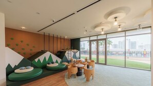 A children's play area with green and white climbing structures, a small table with chairs, and large windows offering a view of palm trees and buildings. - Calm & Stylish 1Bedroom in Hadley Heights JVC (Jumeirah Village Circle (JVC))