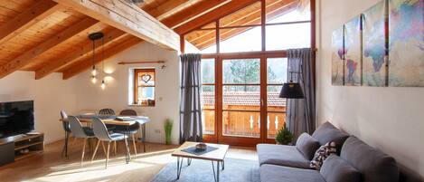 Un salon spacieux avec un plafond en bois, une grande fenêtre, un canapé gris, un coin repas avec des chaises et un téléviseur à écran plat.