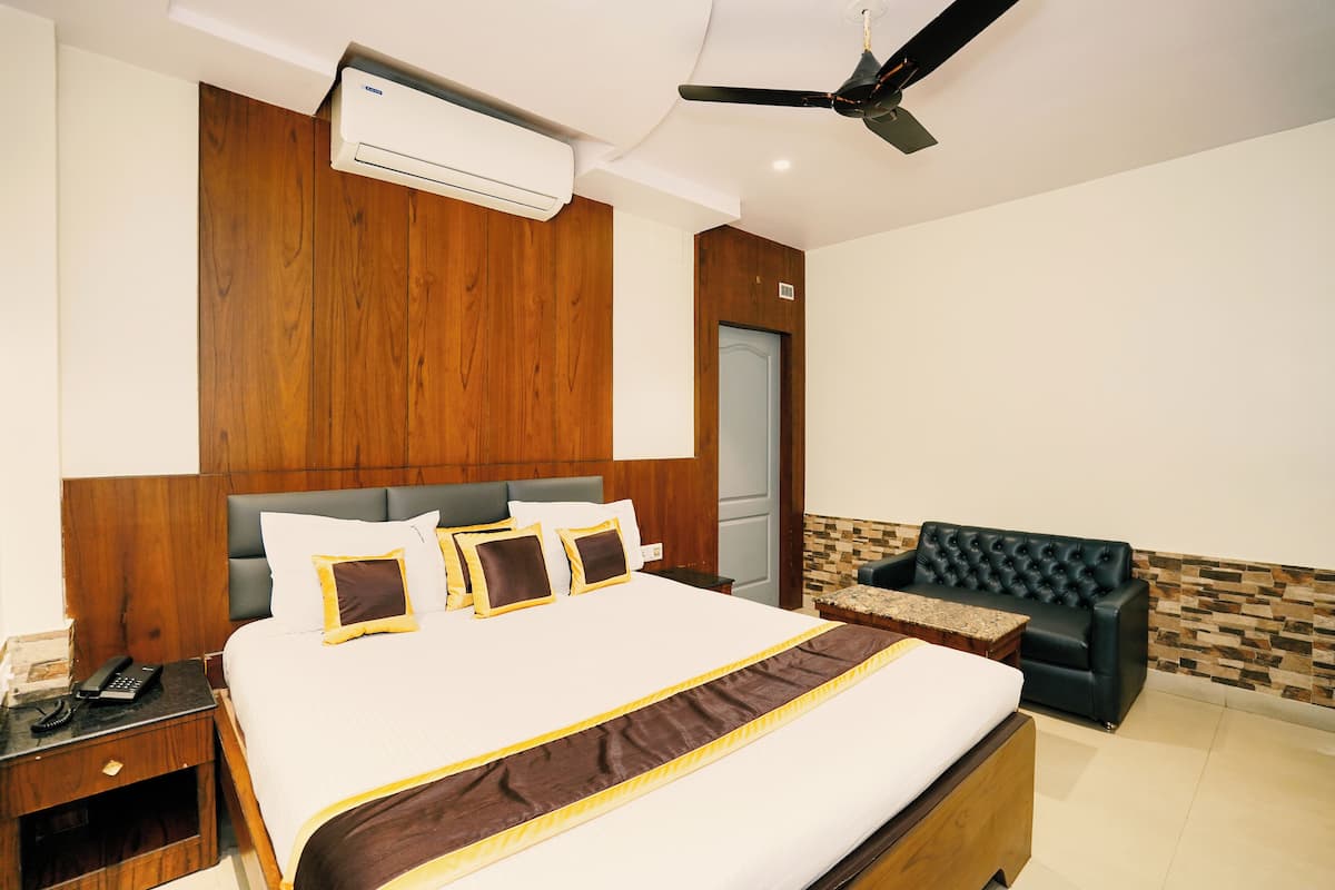 A hotel room with a large bed, a wooden headboard, a ceiling fan, an air conditioning unit, a small table, and a black leather sofa.