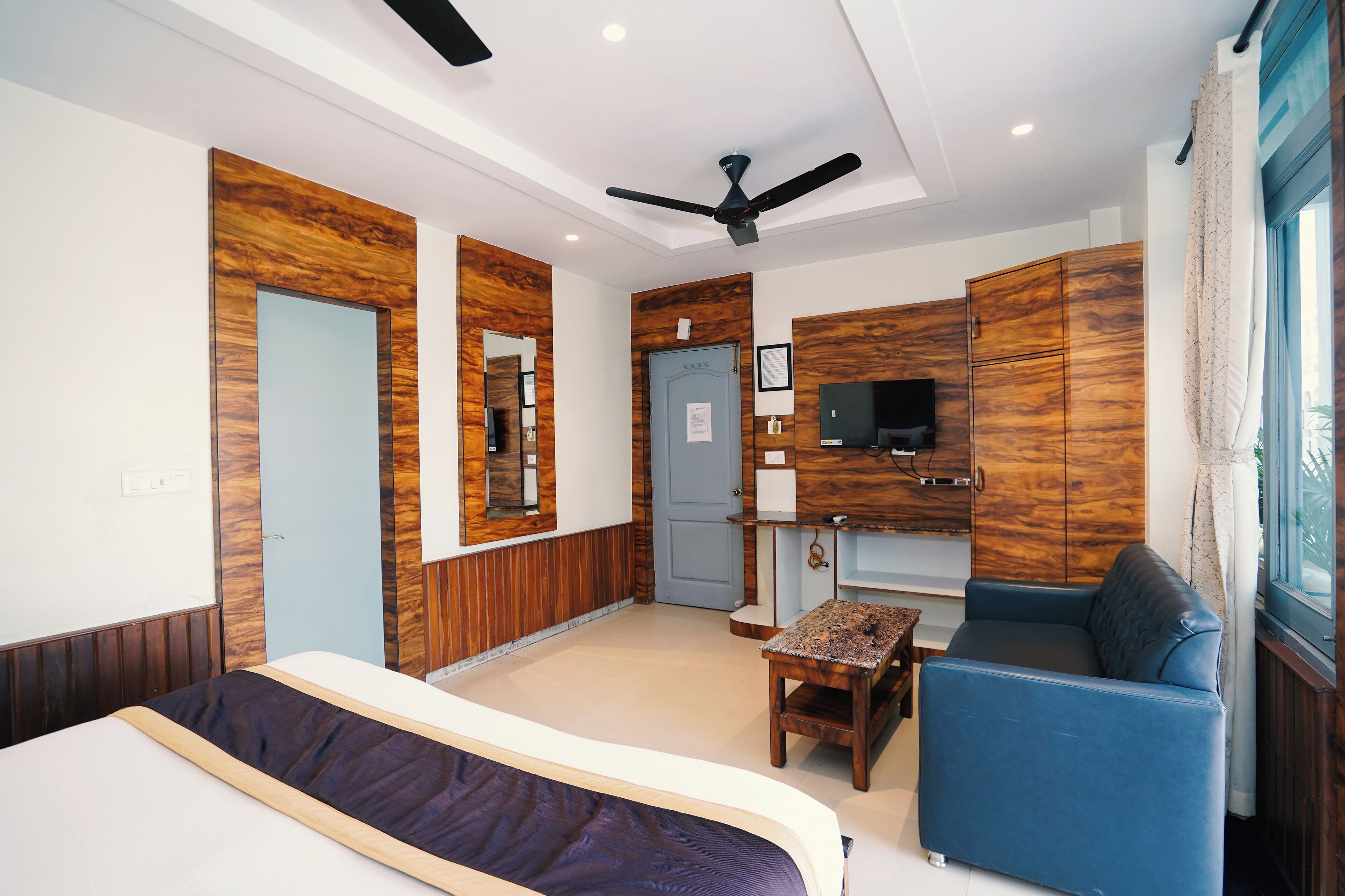 A modern hotel room with a bed, a blue sofa, a small table, a ceiling fan, and wooden paneling on the walls.