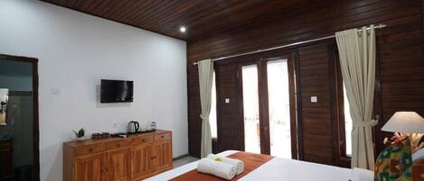 A hotel room with a wooden ceiling, a bed with a white and orange bedspread, a wooden dresser, a flat-screen TV, and a window with curtains.
