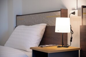 A neatly made bed with white linens, a bedside table with a lamp, and a wall-mounted power outlet.