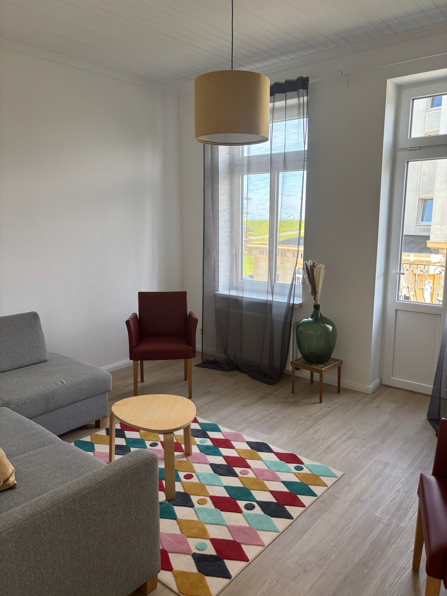 Un salon moderne avec un canapé gris, un fauteuil rouge, une table basse en bois et un tapis coloré. Une grande fenêtre ornée de rideaux légers laisse entrer la lumière naturelle.