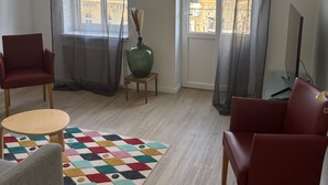 Un salon moderne avec deux fauteuils rouges, une petite table ronde, un tapis coloré, un grand vase et un téléviseur posé sur un meuble.