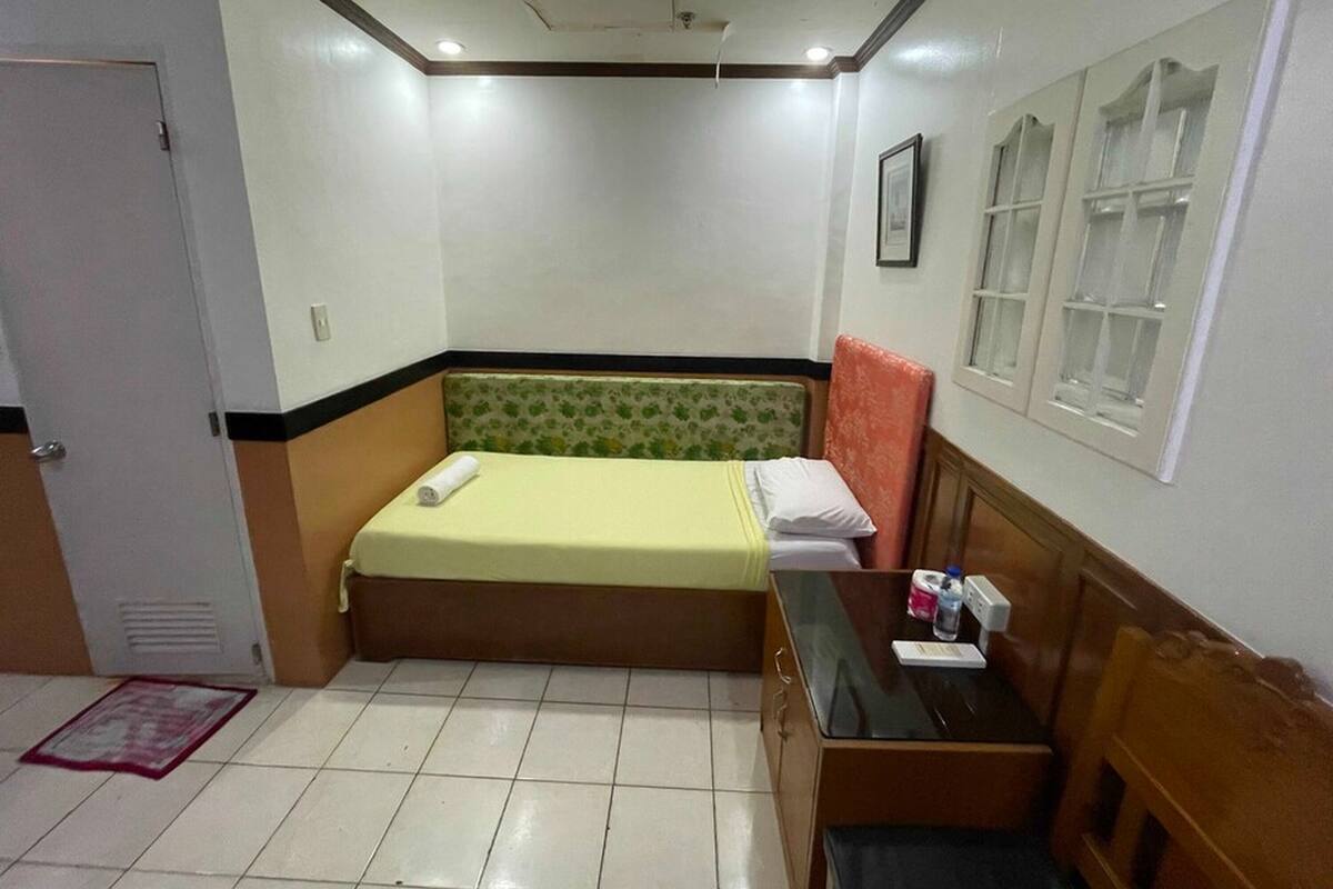 A small, neatly arranged bedroom with a single bed, a bedside table with toiletries, and a window with blinds.