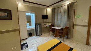 A hotel room with a bed, a desk, a chair, a television, a mirror, a refrigerator, and a window with curtains. - Palette Gems Hotel and Conference Center (Antipolo)