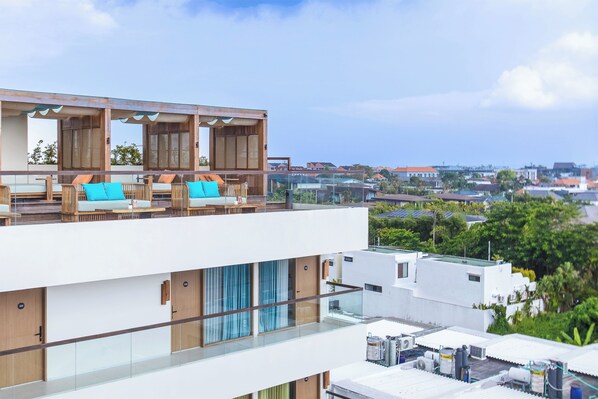 Un immeuble moderne à plusieurs étages, doté d’une terrasse sur le toit aménagée avec des espaces de détente en plein air et offrant une vue sur les environs.