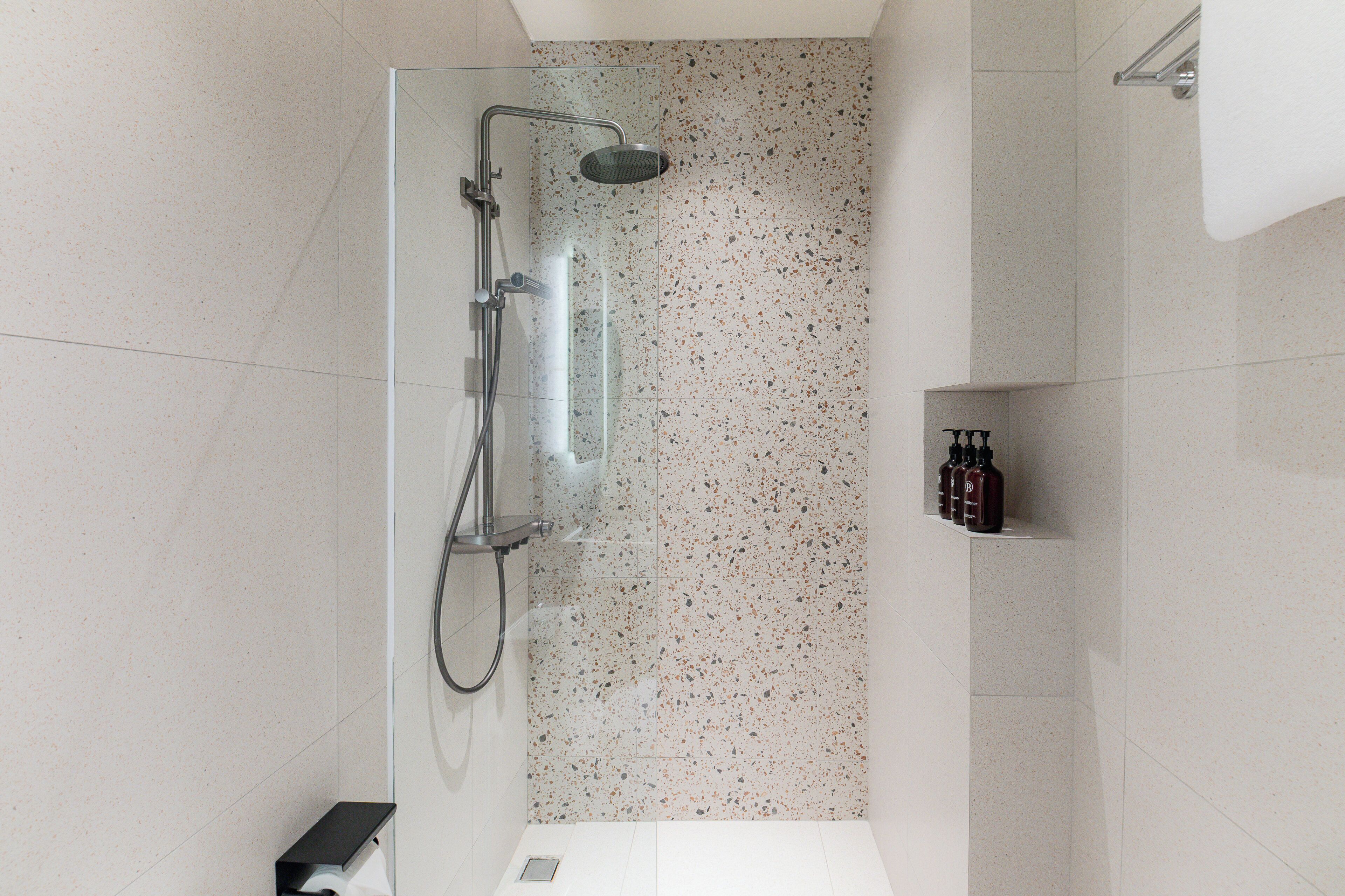 A modern bathroom with a glass-enclosed shower, white speckled tiles, and a small shelf with toiletries.