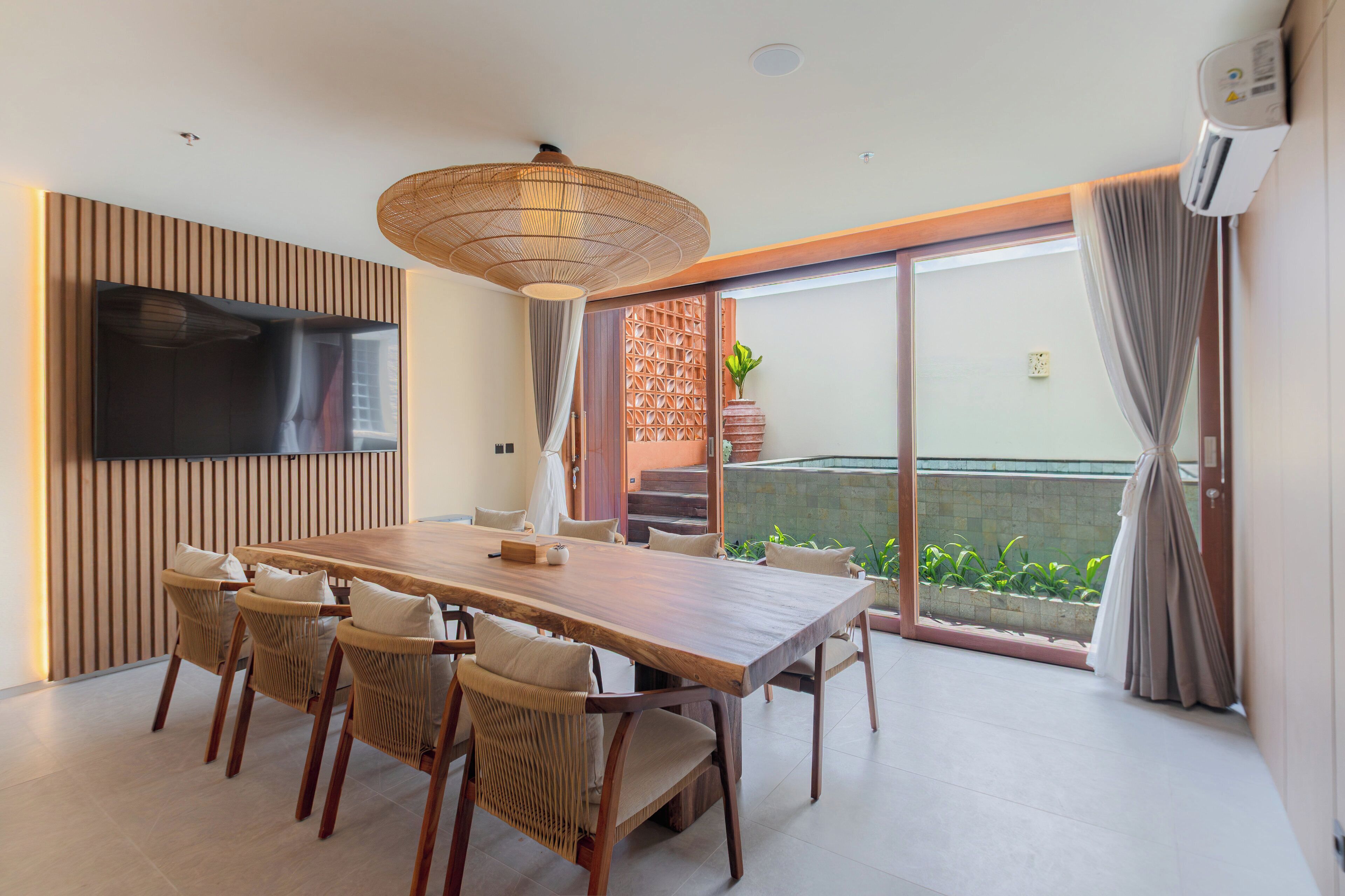 A modern dining area with a wooden table and chairs, a television mounted on the wall, and a sliding glass door leading to a balcony with plants.