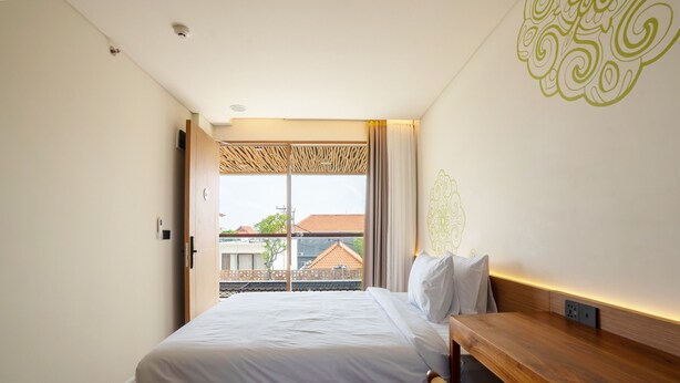 Habitación de hotel con cama, un banco de madera, ventana con vistas a edificios y un diseño decorativo en la pared.