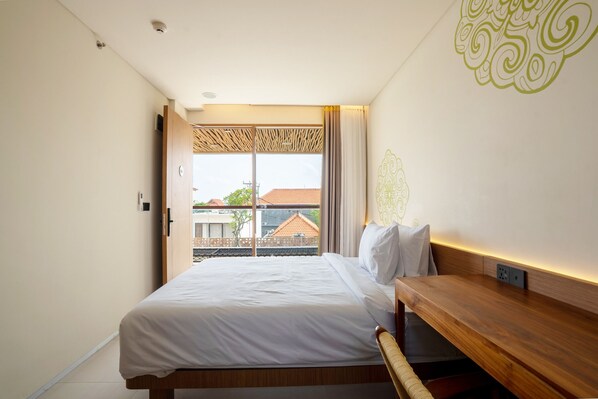 A hotel room with a bed, a wooden bench, a window with a view of buildings, and a decorative wall pattern. - BLive Coliving Canggu (Canggu)