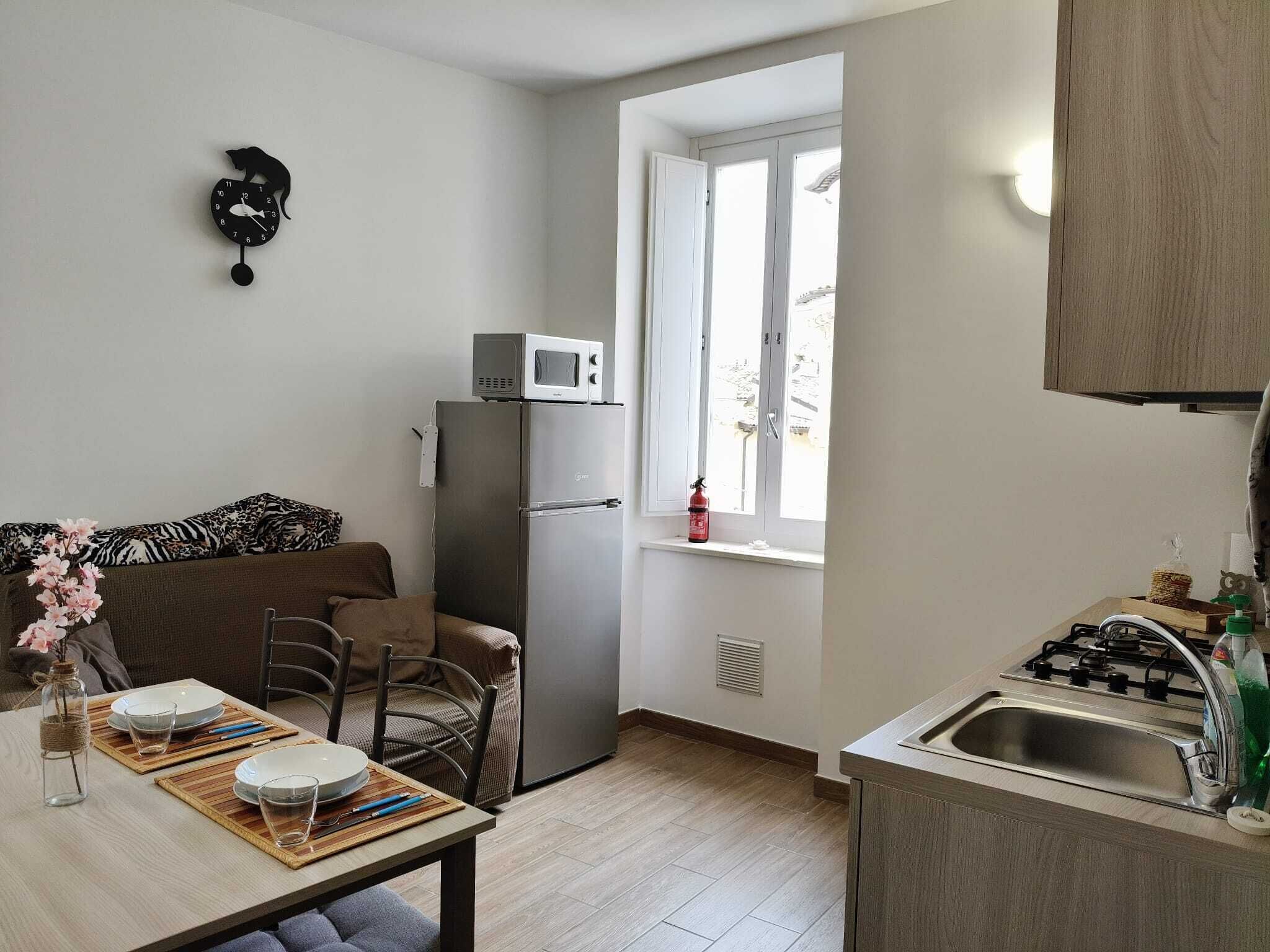 A compact kitchen with a microwave, a refrigerator, a window with a view, a dining area with a table set for two, and a small sink.
