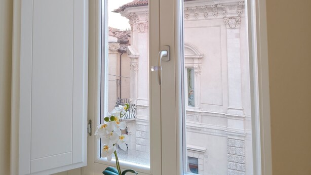 A room with a large window, a white vase with flowers on the windowsill, and a view of a building with classical architecture. - Design and History in the Heart of L'Aquila Capital of Culture 2026 (L'Aquila)