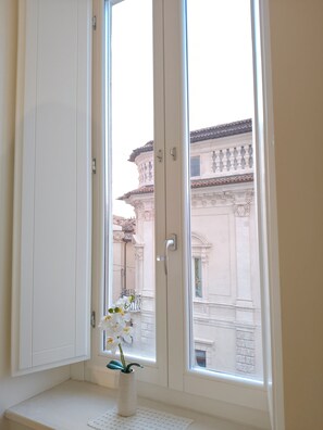 Une pièce dotée d’une grande fenêtre, d’un vase blanc orné de fleurs posé sur le rebord de la fenêtre, et offrant une vue sur un bâtiment à l’architecture classique.