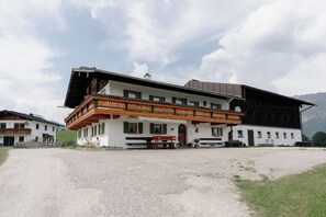A traditional chalet-style building with a balcony, a gravel driveway, and mountainous background. - Sleeping Witch, 50 m², sleeps 1–4, 2 bedrooms, shower/toilet (Bischofswiesen)