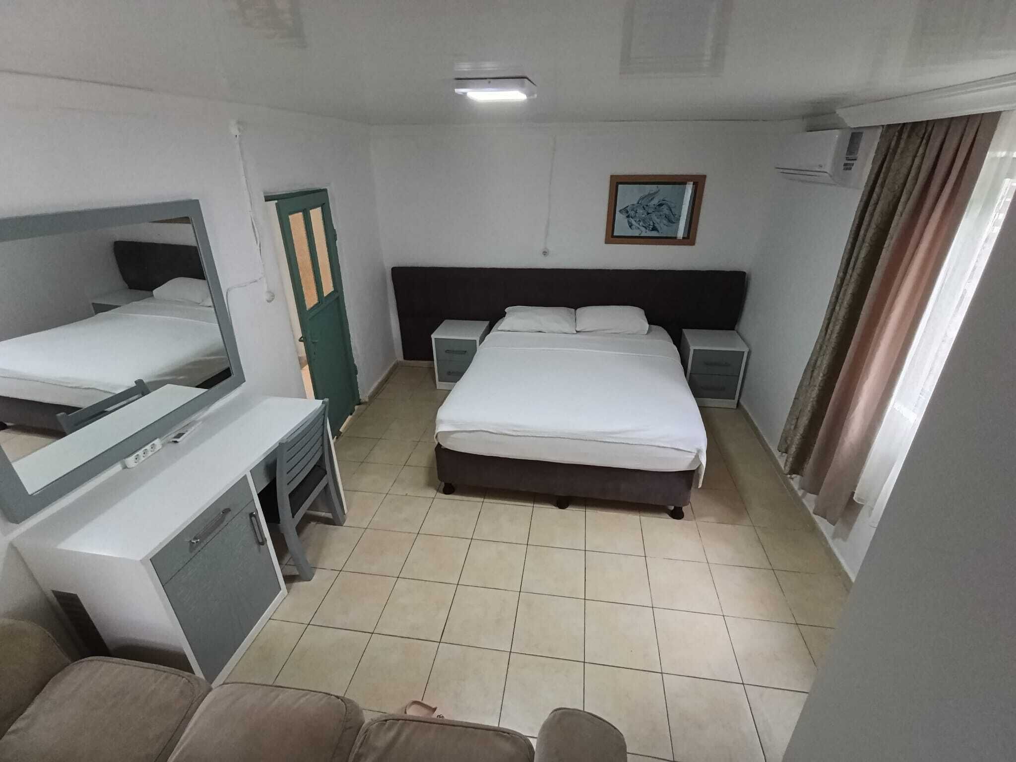 A hotel room with a bed, a desk, a chair, a wardrobe, a mirror, a nightstand, a bedside table, and a window with curtains.