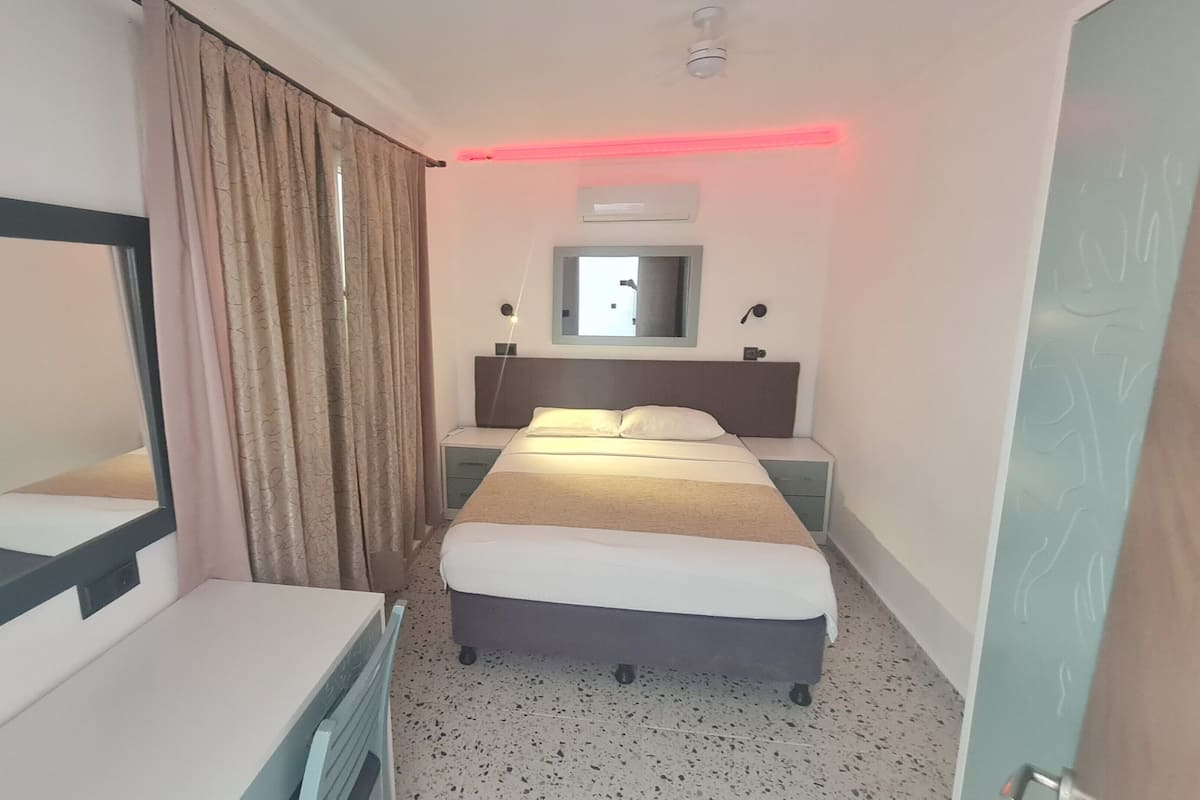 A hotel room with a bed, a bedside table, a mirror, a window with curtains, an air conditioning unit, and a ceiling fan.