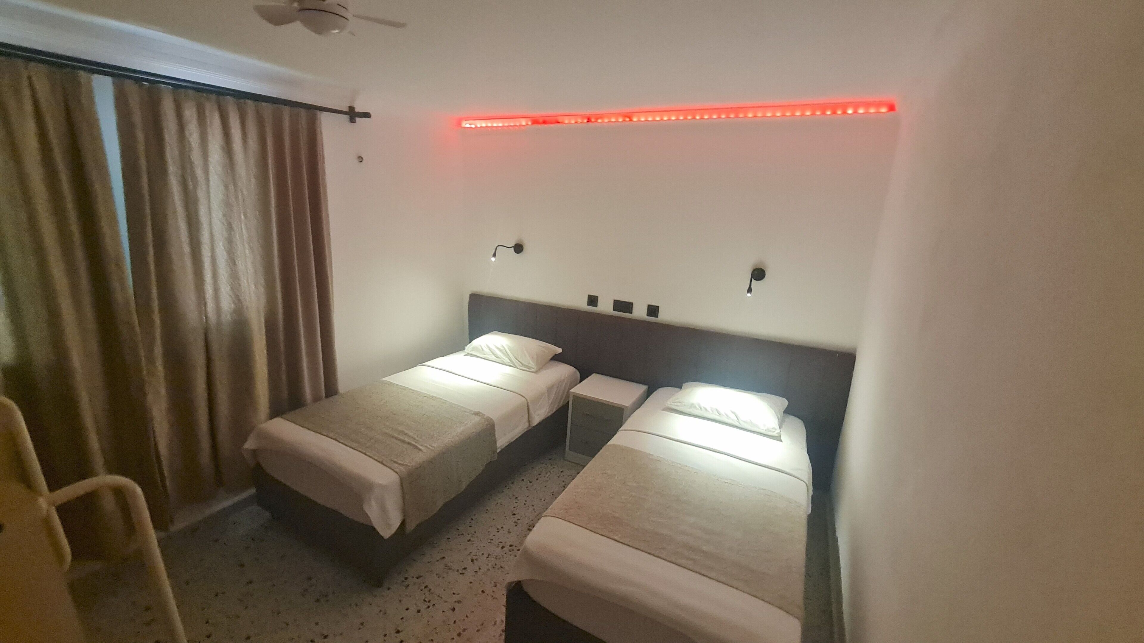 Habitación de hotel con dos camas individuales, ventilador de techo, tira de luces LED rojas y ventana con cortinas.