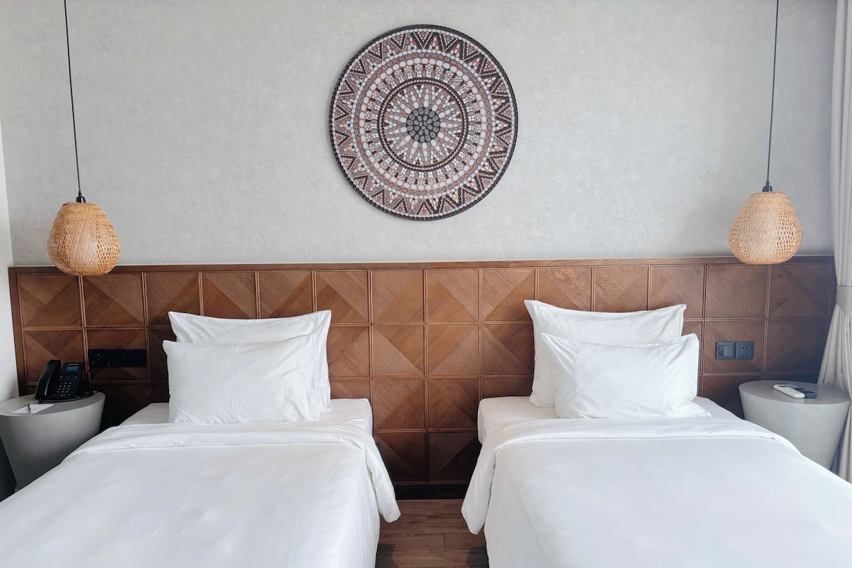 A hotel room with two beds, a patterned wall hanging, and two wicker lamps.