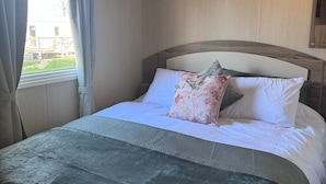 Cama bem arrumada com almofada florida e manta cinza, janela com cortinas e cabeceira de madeira.