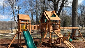 Eine hölzerne Spielkonstruktion mit Rutsche und Kletterelementen, umgeben von Bäumen und blauem Himmel.