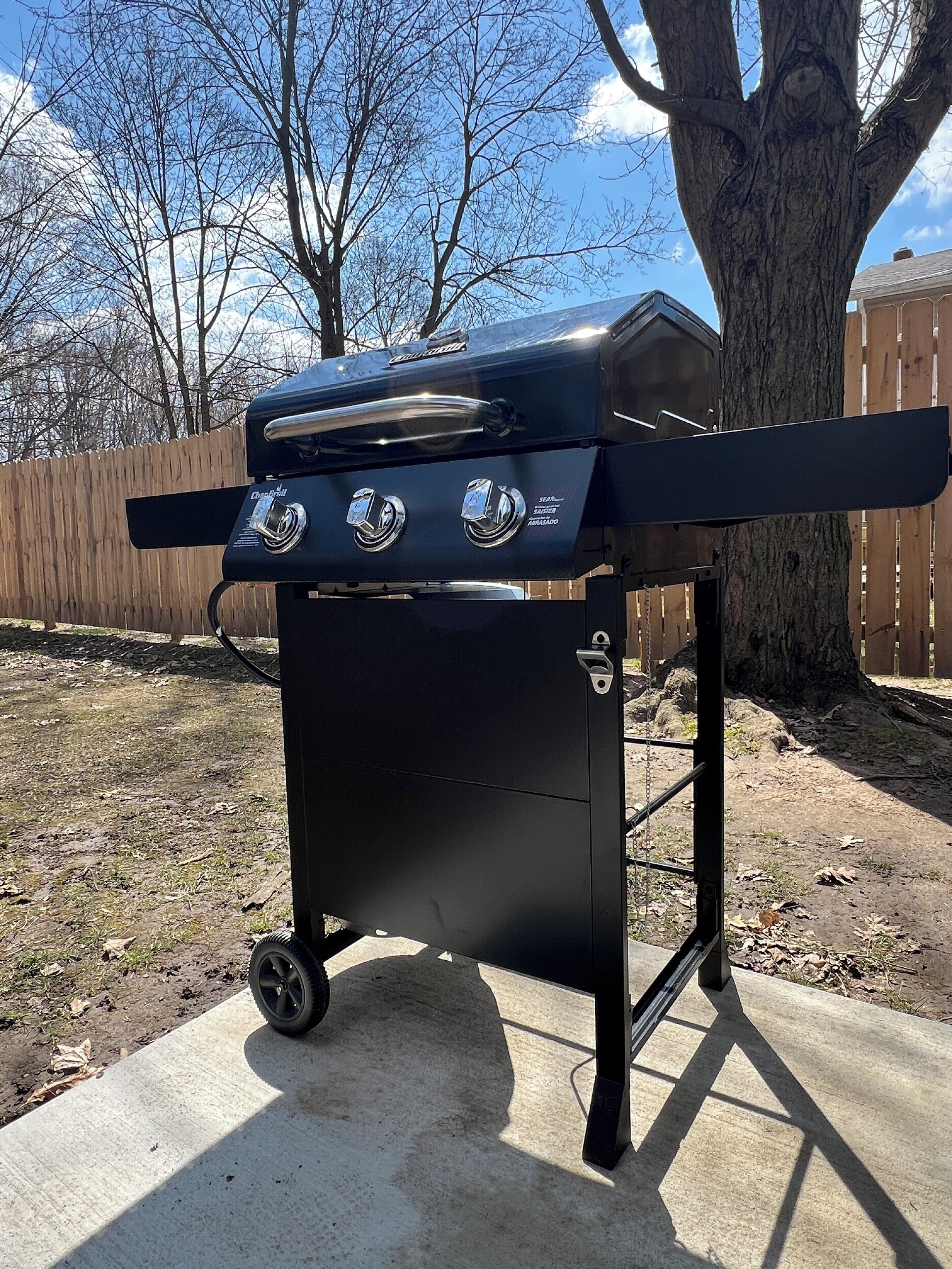 En portabel grill med två brännare och en sidohylla, placerad på en betongterrass med ett trästaket och träd i bakgrunden.