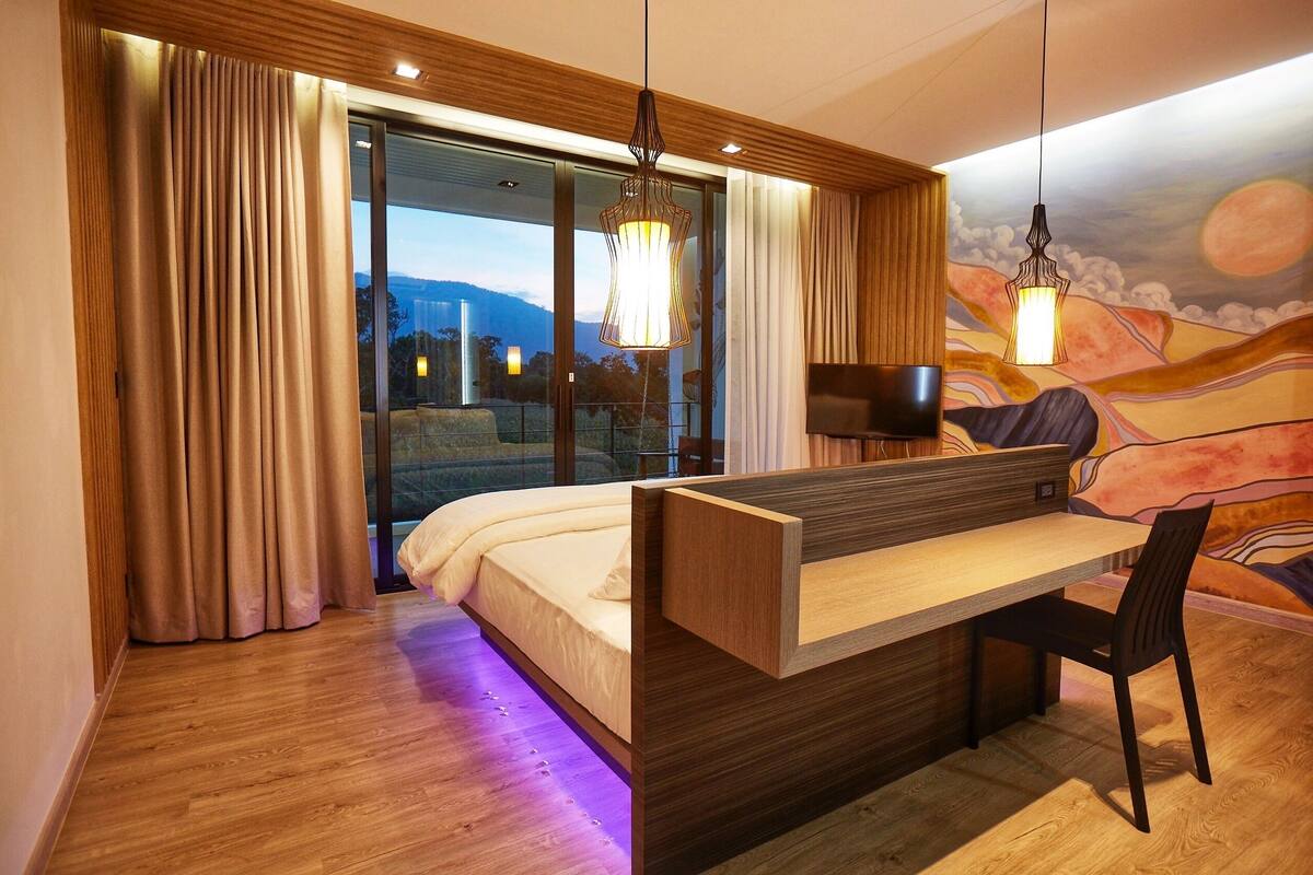 Un dormitorio moderno con cama, escritorio y silla, ventanas amplias y un mural en la pared.