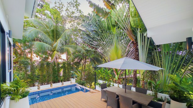 Zona junto a la piscina con terraza de madera, mobiliario exterior y exuberante vegetación tropical.