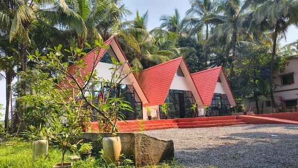 A building with a red roof and white walls, surrounded by palm trees and greenery. - Rustic Palms - Restaurant & Cabins (Alibag)