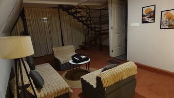 Una sala de estar con sofá, sillón y una mesita. Hay una lámpara, una escalera y una puerta que da a otra habitación.