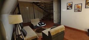 A living room with a sofa, armchair, and a small table. There is a lamp, a staircase, and a door leading to another room.
