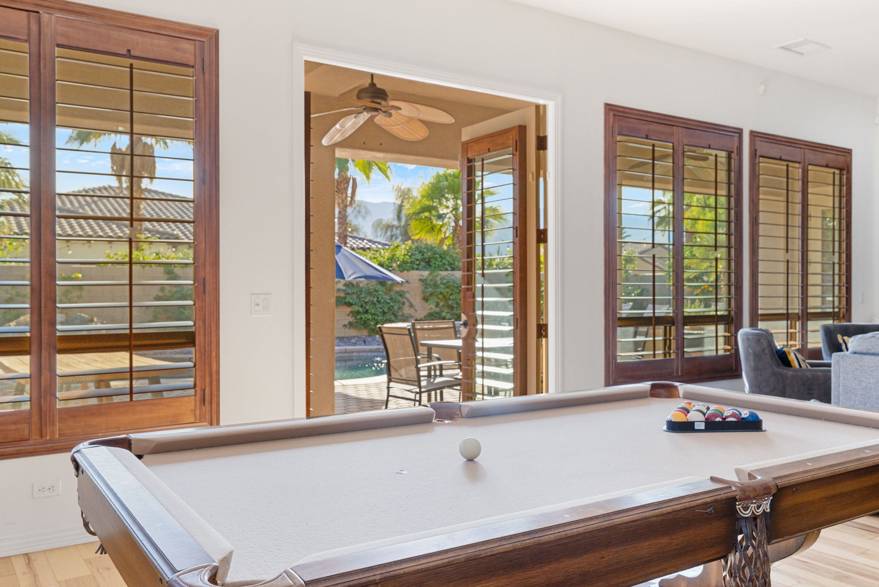 A pool table in a room with large wooden-framed windows, a ceiling fan, and a view of an outdoor patio with a table and chairs.