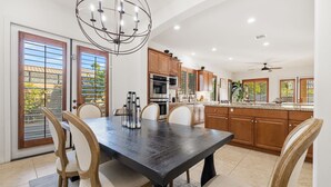 A modern kitchen with wooden cabinets, a central island, and a dining area with a dark table and light chairs. - Relaxing Vacay Pool Spa Fire Pit Family Games (Indio)