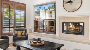 A living room with a fireplace, a large window with a view of palm trees, and a ceiling fan. - Relaxing Vacay Pool Spa Fire Pit Family Games (Indio)