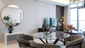 A modern living room with a round glass table, four grey upholstered chairs, a large round mirror, a flat-screen TV, and a decorative geometric sculpture. - Elegant 1BR Apartment, Rooftop 360° Infinity Pool with Burj Views (Dubai)